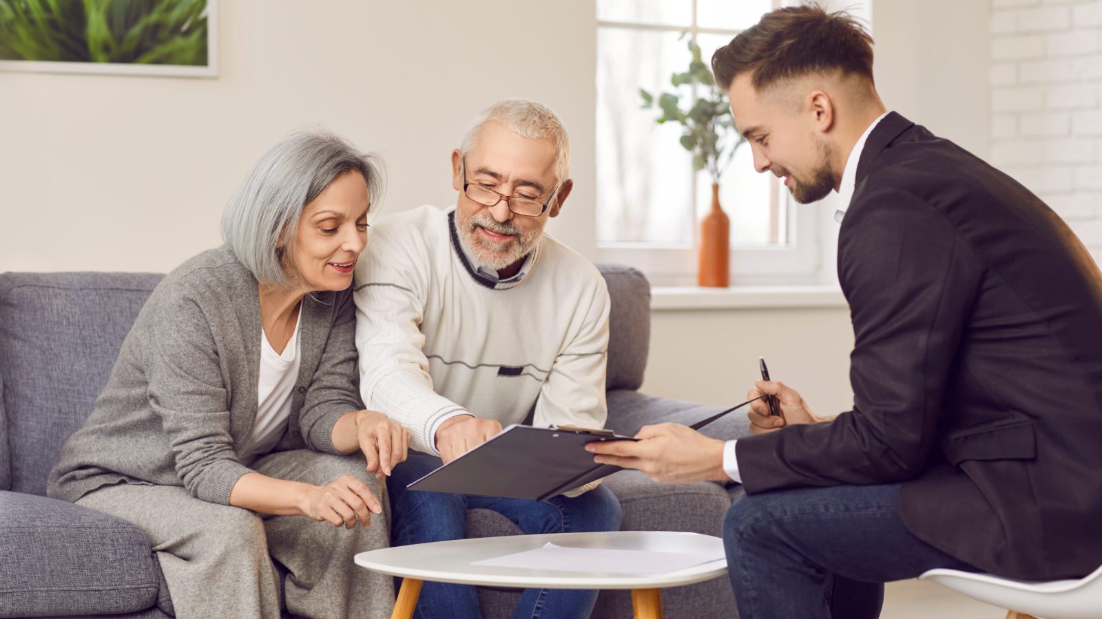 Elderly couple is carefully studying documents provided by financial advisor in cozy living room. Smiling mature family discussing retirement planning, property arrangements or investment choices.