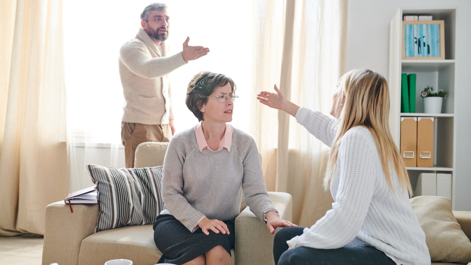 Frowning family psychologist in glasses sitting in armchair and listening to quarrel of couple while asking young lady to stop conflict, emotional couple gesturing hands and blaming each other for their problems
