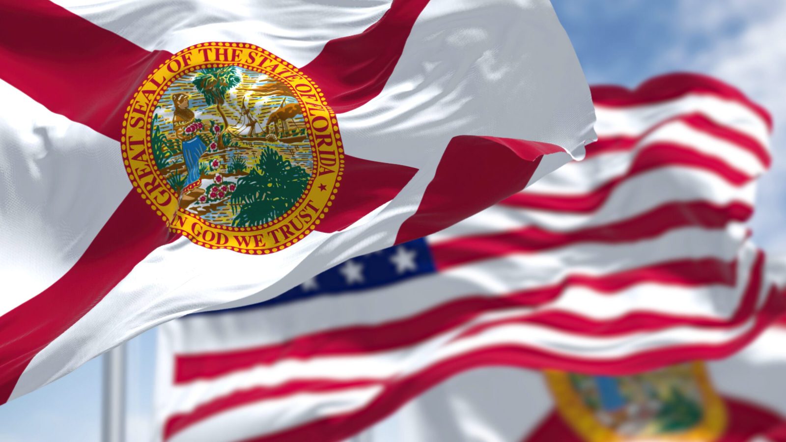 The Florida state flag waving along with the national flag of the United States of America. In the background there is a clear sky.