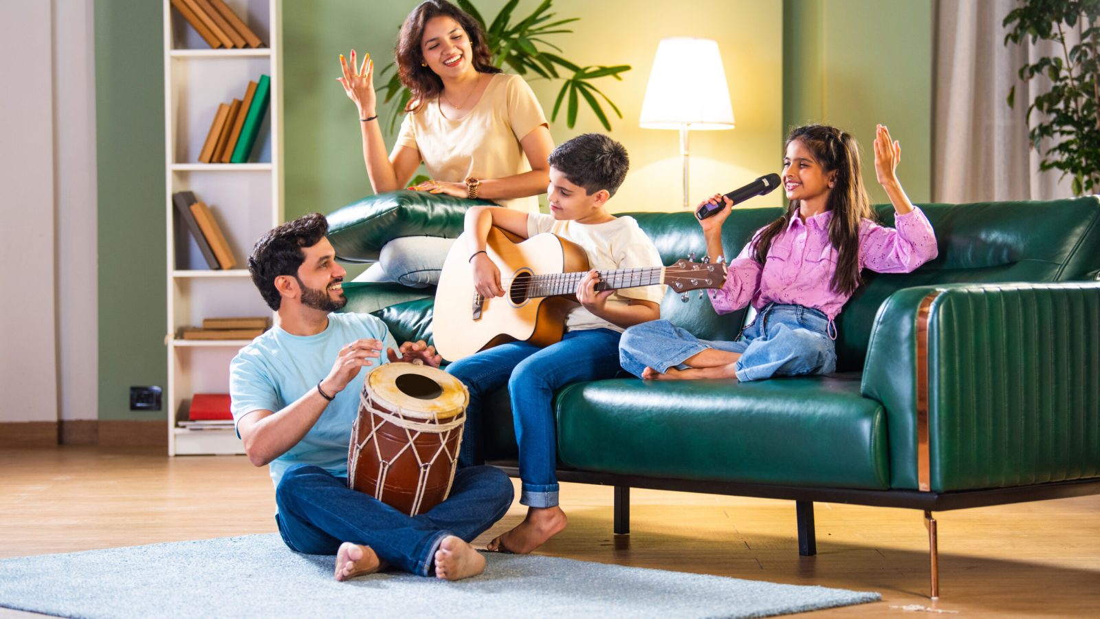 Joyful Indian Asian family enjoying music at home: daughter singing, son on guitar, father on dholki, mother appreciating, celebrating togetherness on living room sofa
